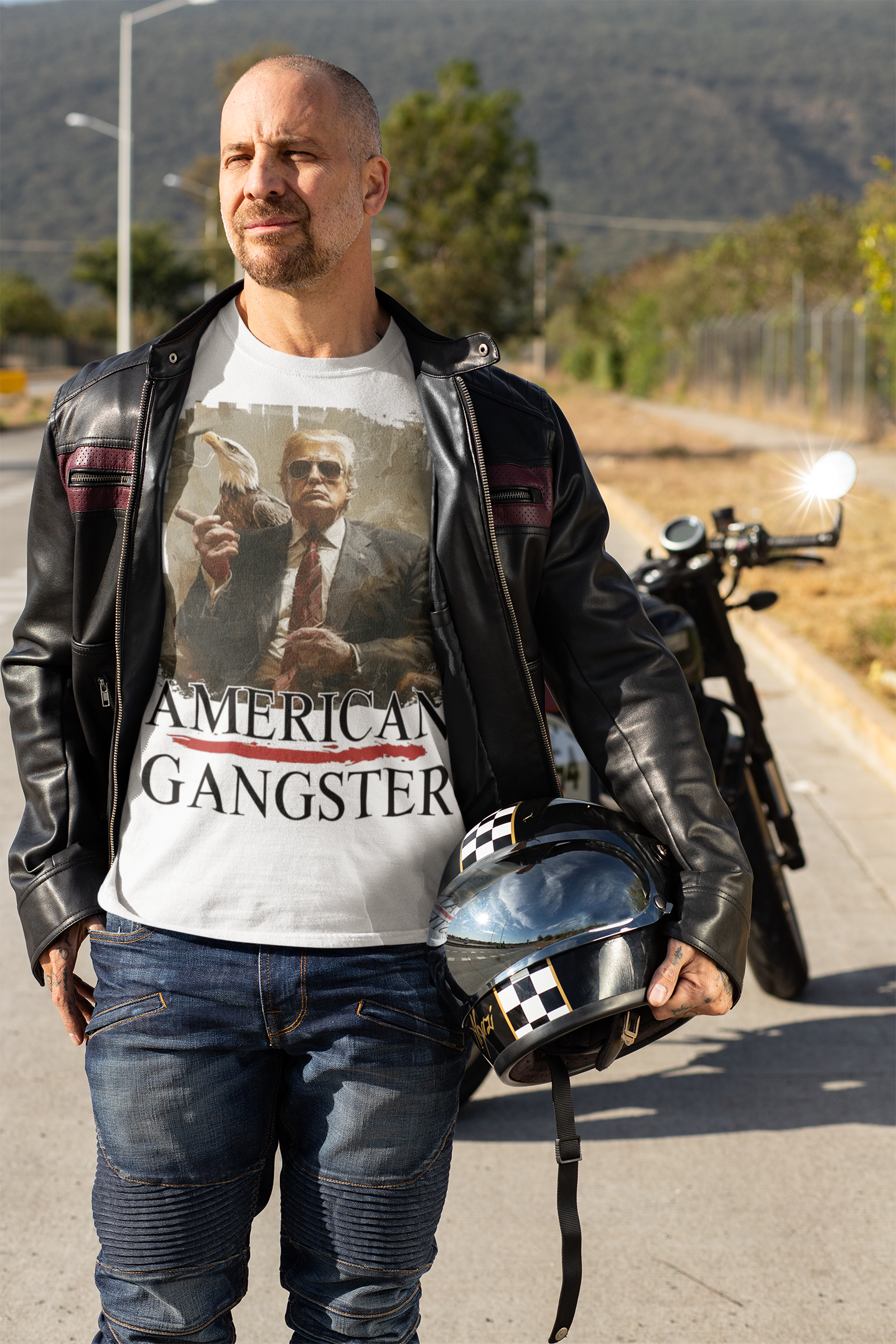 Man wearing American Gangster Graphic Design t-shirt, standing next to a motorcycle on a scenic road.