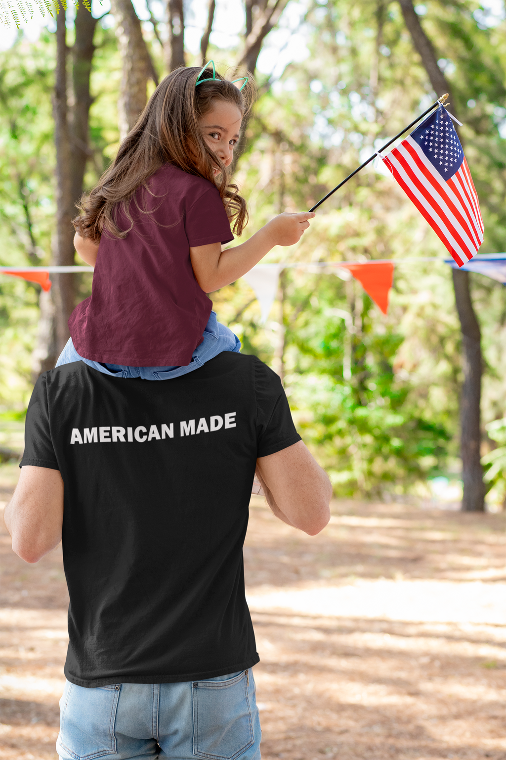 Father carrying daughter with American flag, showcasing Authentic American Made Apparel in a festive outdoor setting.