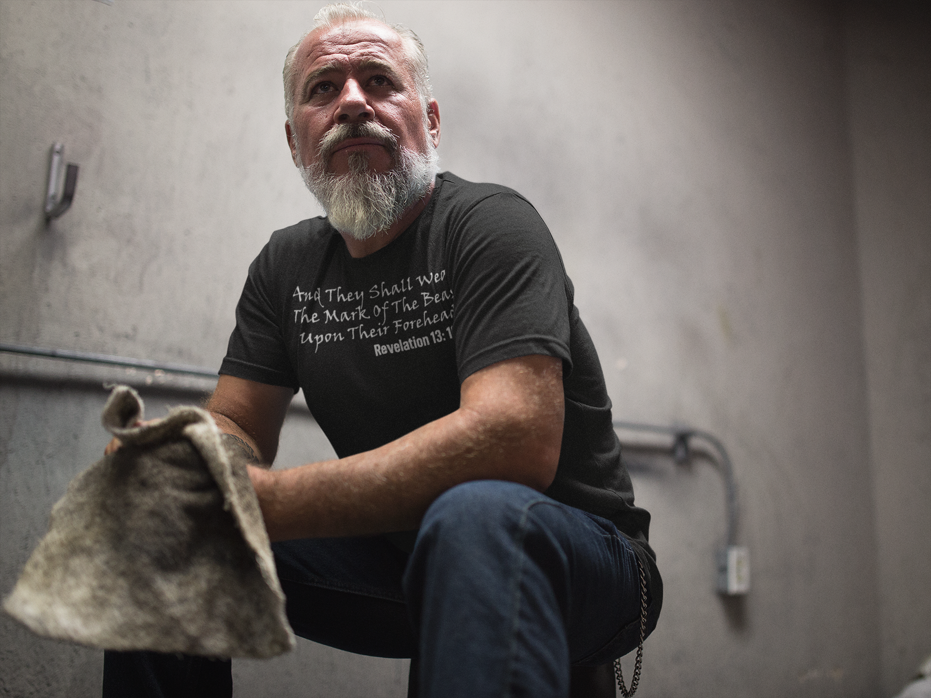 Man wearing The Mark Of The Beast Revealed graphic tee, seated and thoughtful in a dimly lit space.