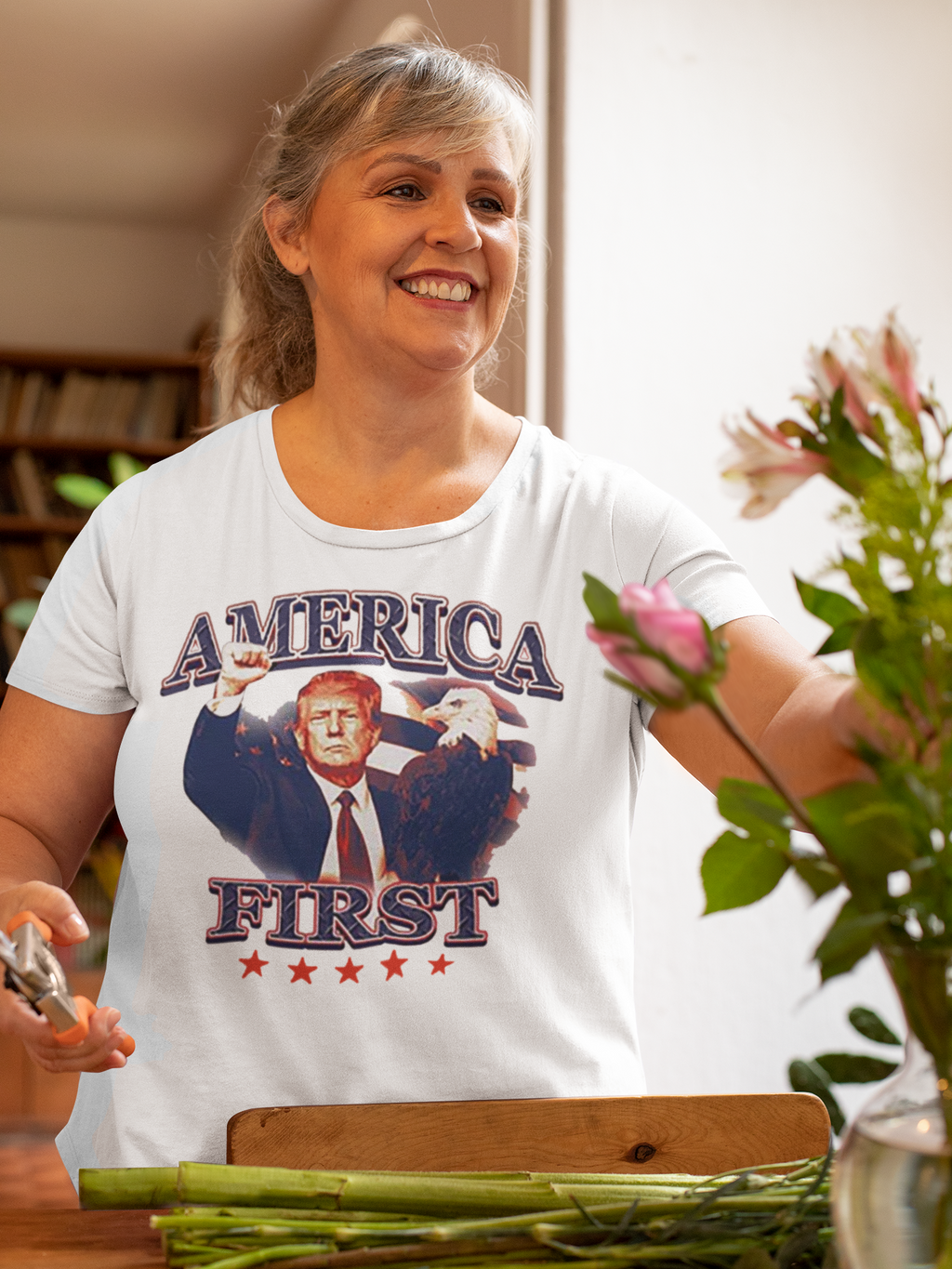 Woman wearing America First Graphic Design Tee while arranging flowers in a cozy indoor setting.