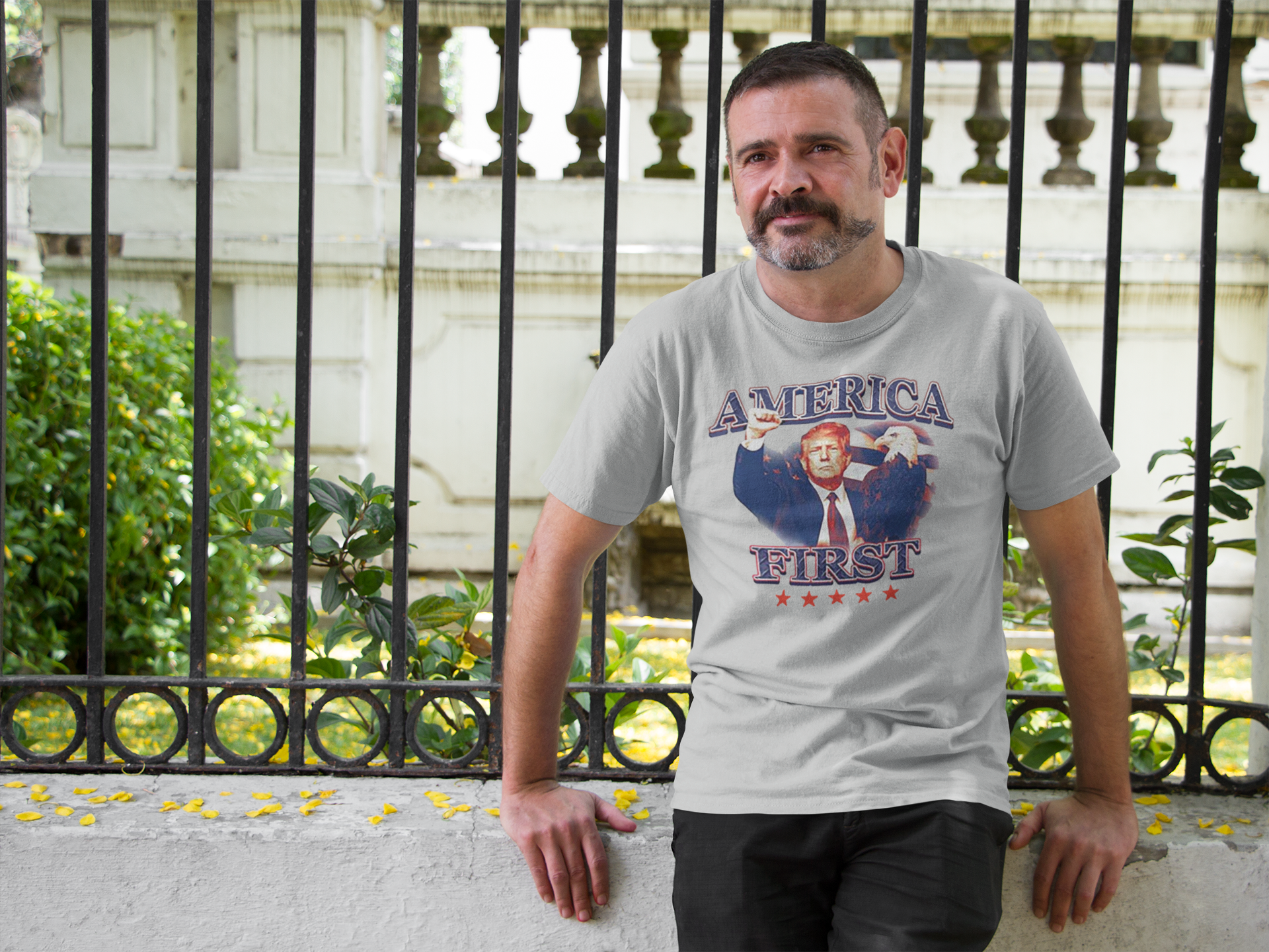 Man wearing America First Graphic Design Tee outdoors by a decorative fence.