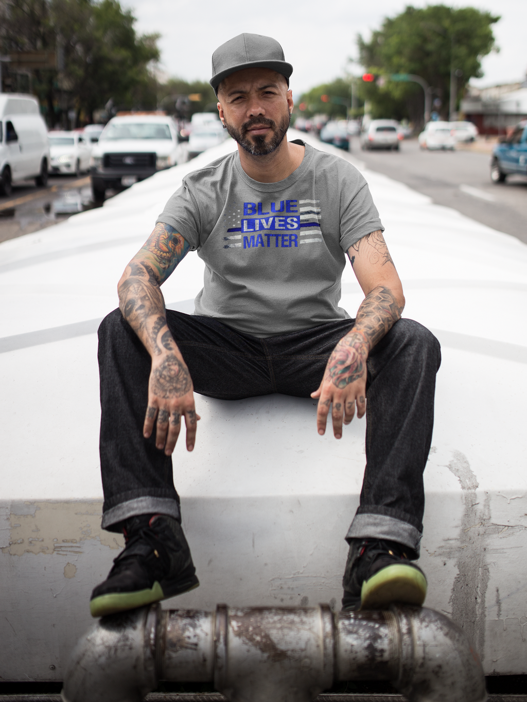 Man wearing Blue Lives Matter American Flag Patriotic Graphic Tee sitting outdoors on a city street structure.