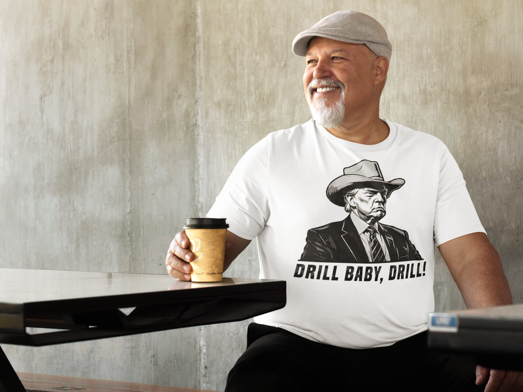 Man wearing Drill Baby Drill Patriotic Graphic Tee while holding coffee, seated at a table with a relaxed smile.