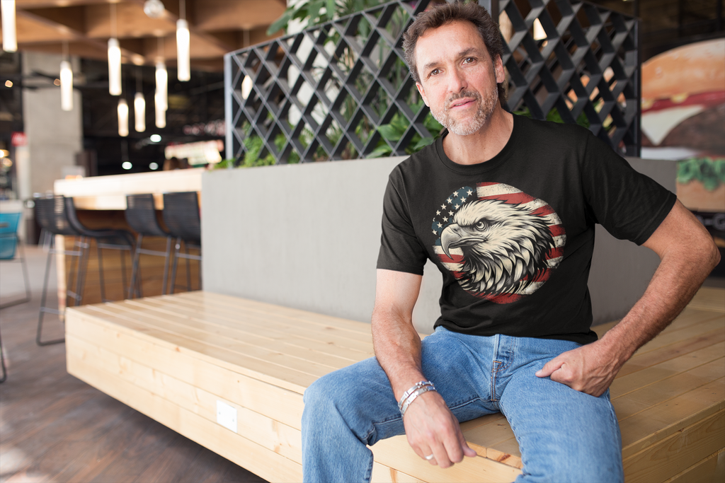 Eagle On American Flag Gear Tee worn by a man sitting in a modern café setting.