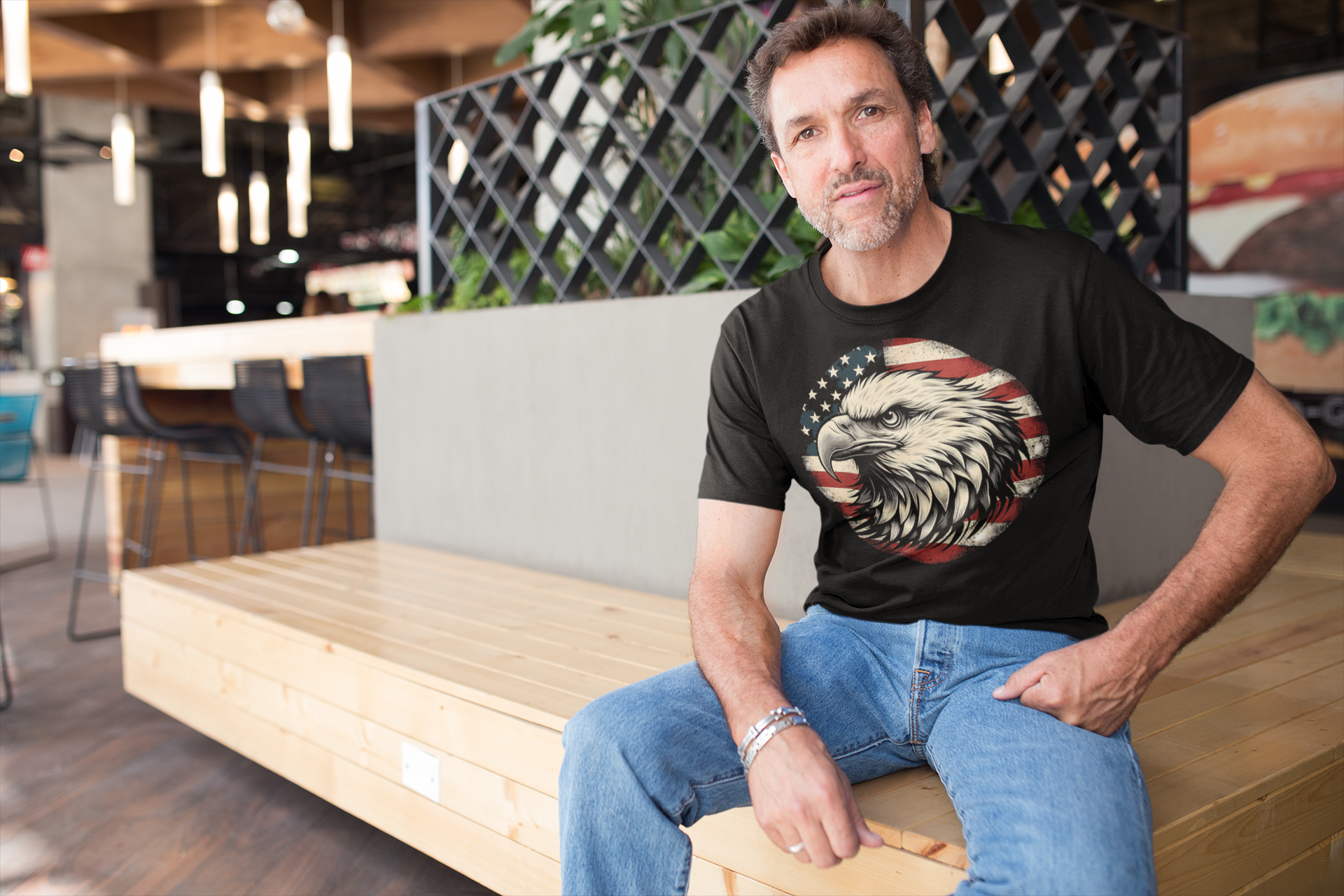 Eagle On American Flag Gear Tee worn by a man sitting in a modern café setting.