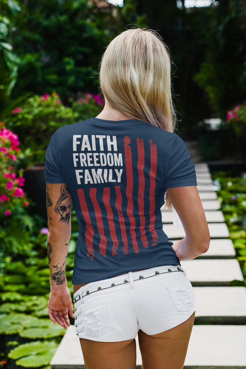A woman wearing a shirt featuring the design 'Faith Freedom Family Image On Back' in a garden setting.
