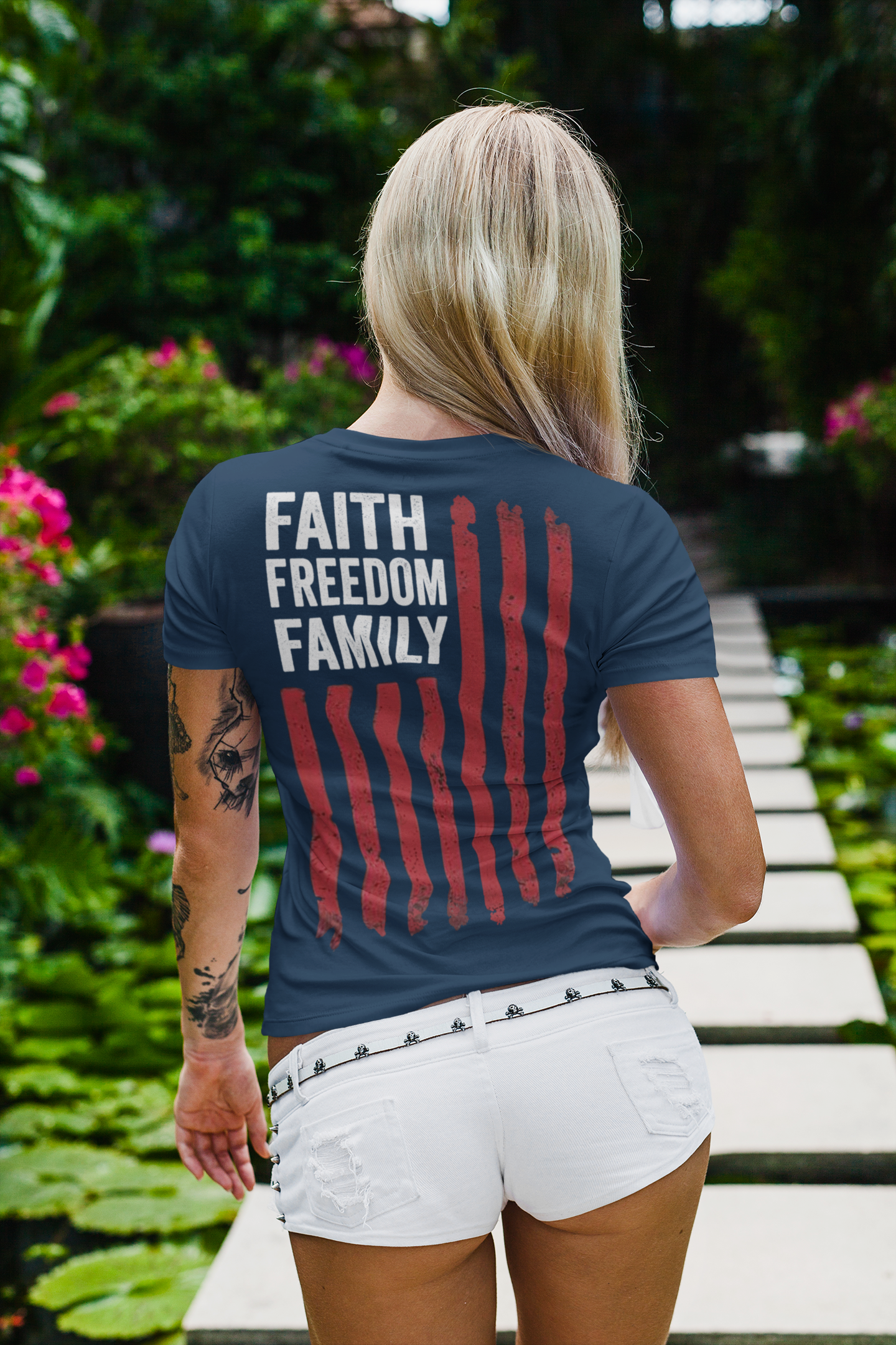 A woman wearing a shirt featuring the design 'Faith Freedom Family Image On Back' in a garden setting.