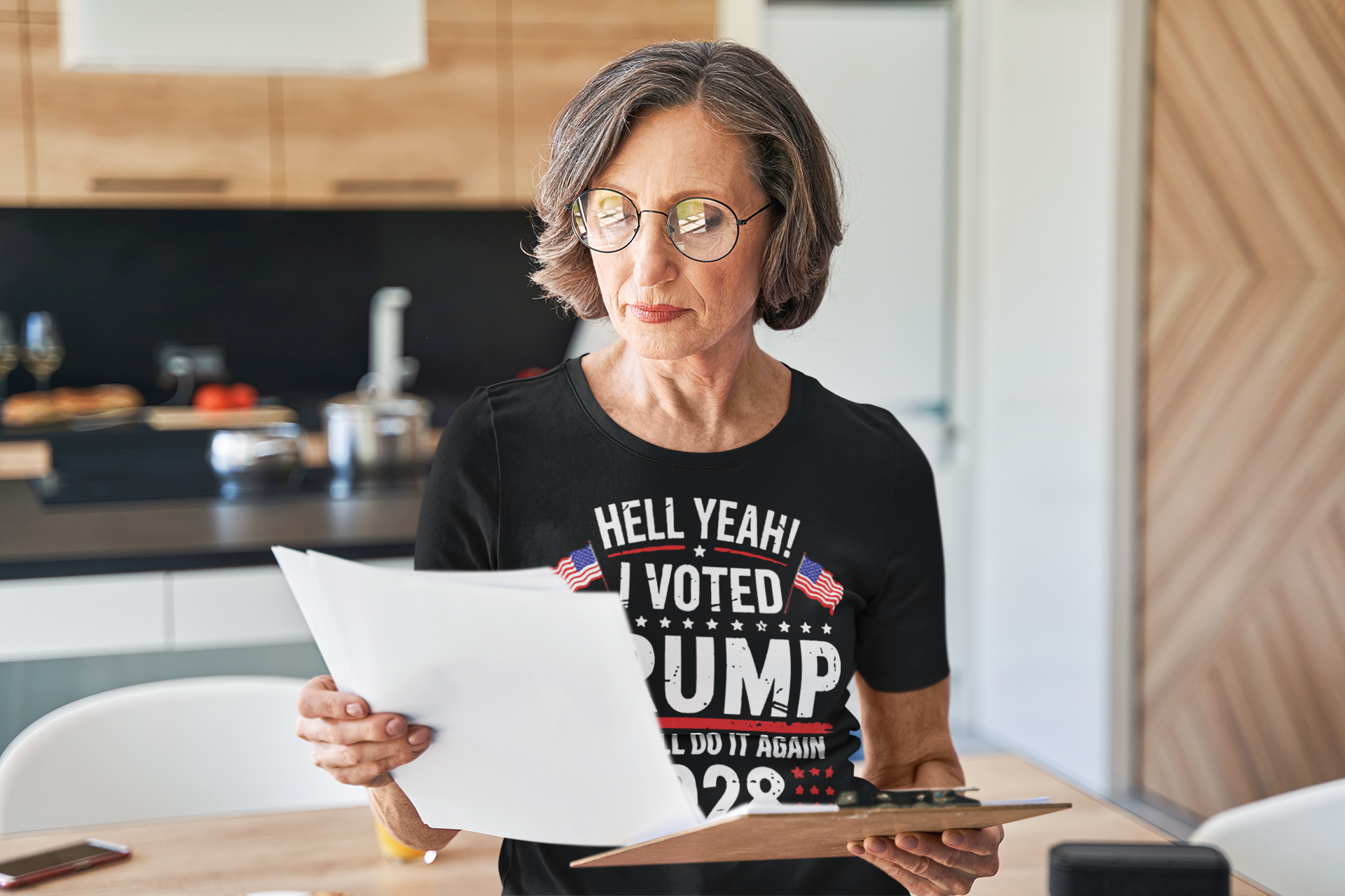 Older woman wearing a shirt that says Hell Yeah I Voted Again 2028 Trump, holding documents in a modern kitchen.
