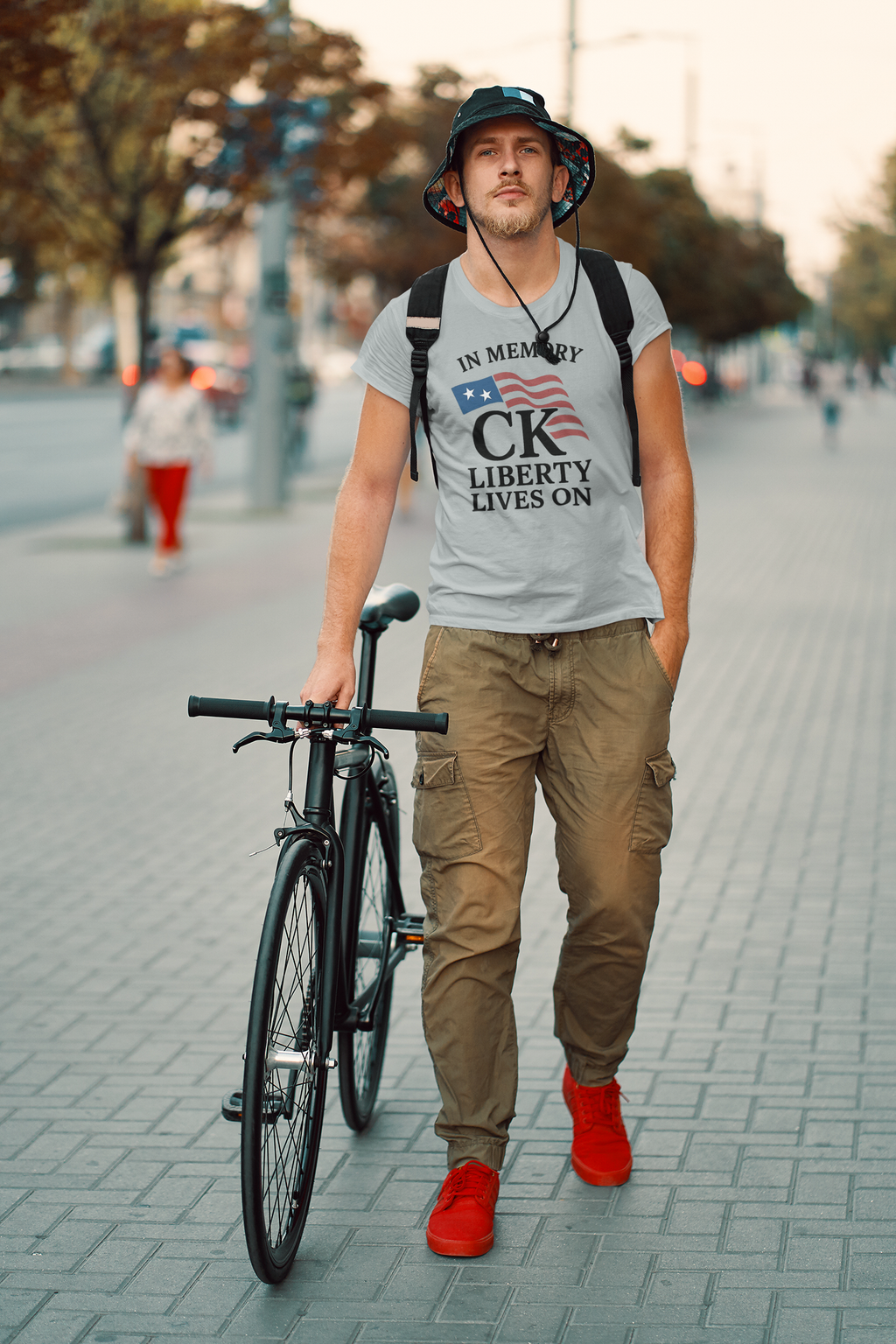 In Memory Of CK Charlie Kirk Liberty Lives On apparel worn by a man walking with a bicycle on a city street.