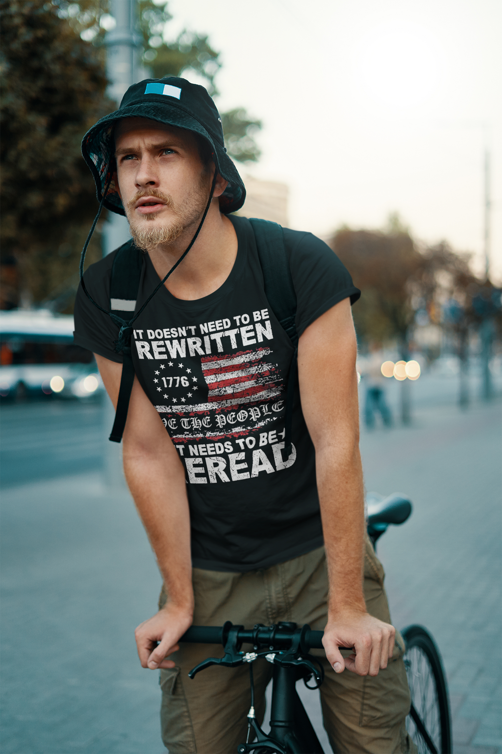 Man in trendy bucket hat and t-shirt with 'It Needs to Be Reread Patriotic Design' riding a bicycle in urban setting.