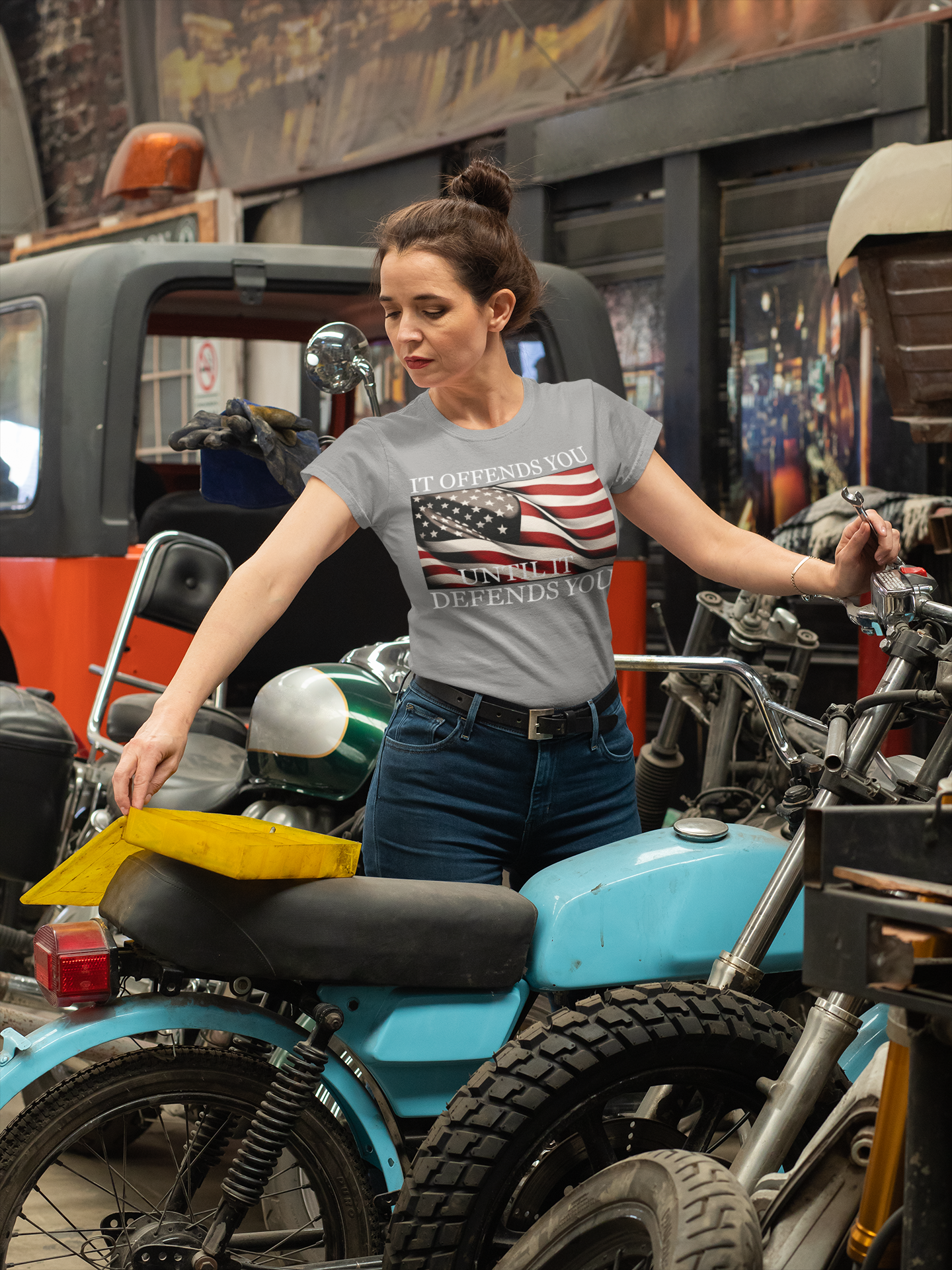 Woman wearing "It Offends You Until It Defends You Wht Ltrs" shirt in a garage with motorcycles.