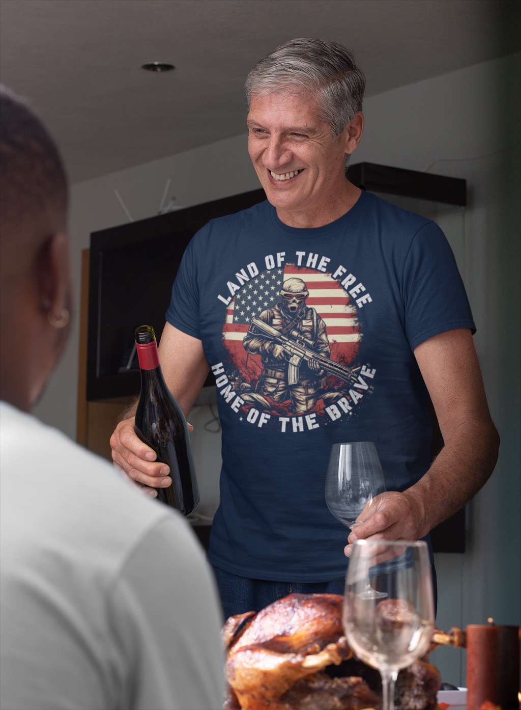 A man smiling while wearing a t-shirt that says Land Of The Free Home Of The Brave, holding a wine bottle and glass.