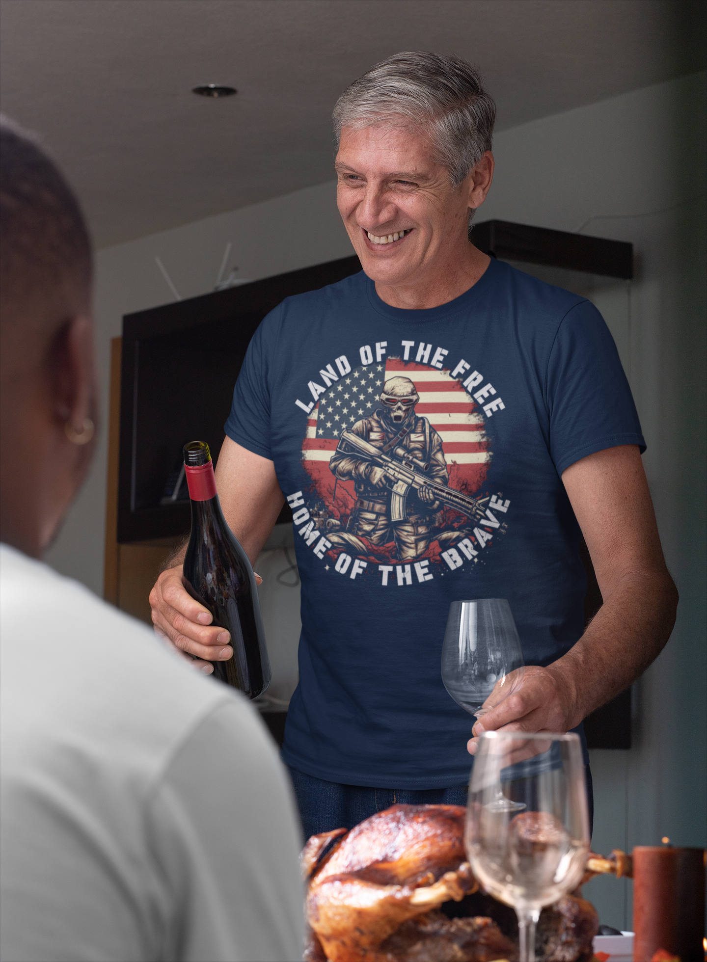 A man smiling while wearing a t-shirt that says Land Of The Free Home Of The Brave, holding a wine bottle and glass.