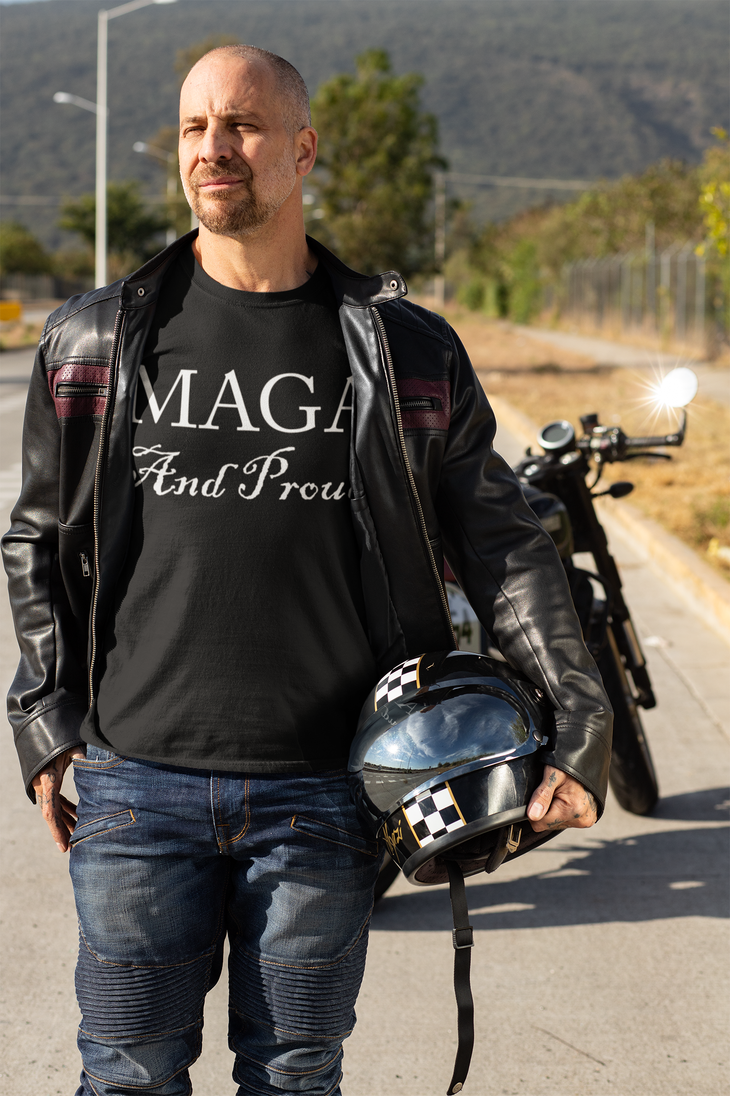 Man wearing a black MAGA And Proud White Letters Style t-shirt and holding a helmet next to a motorcycle.