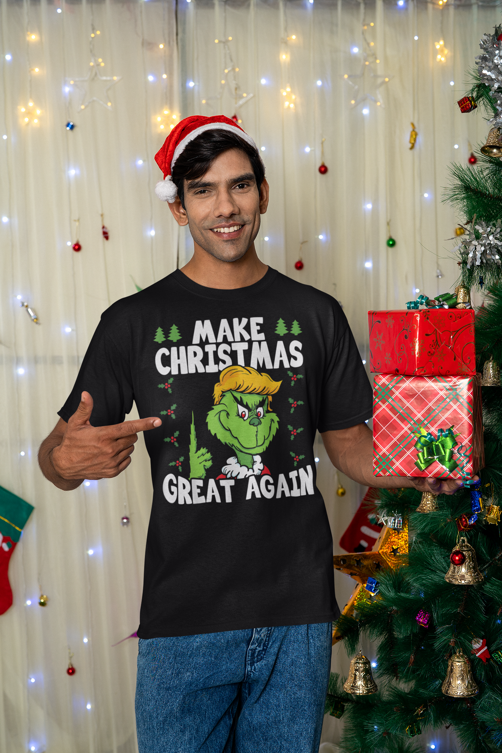Make Christmas Great Again With The Grinch graphic t-shirt worn by a man holding holiday gifts in front of a Christmas tree.