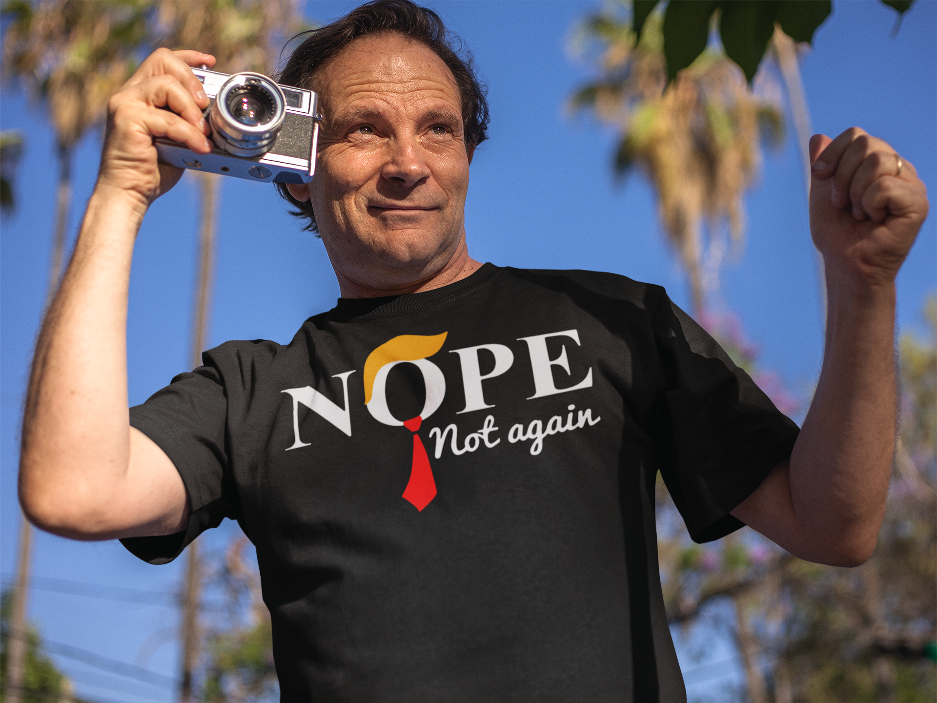 Man wearing Nope Not Again Trump Hair & Tie graphic tee holding a camera outdoors.