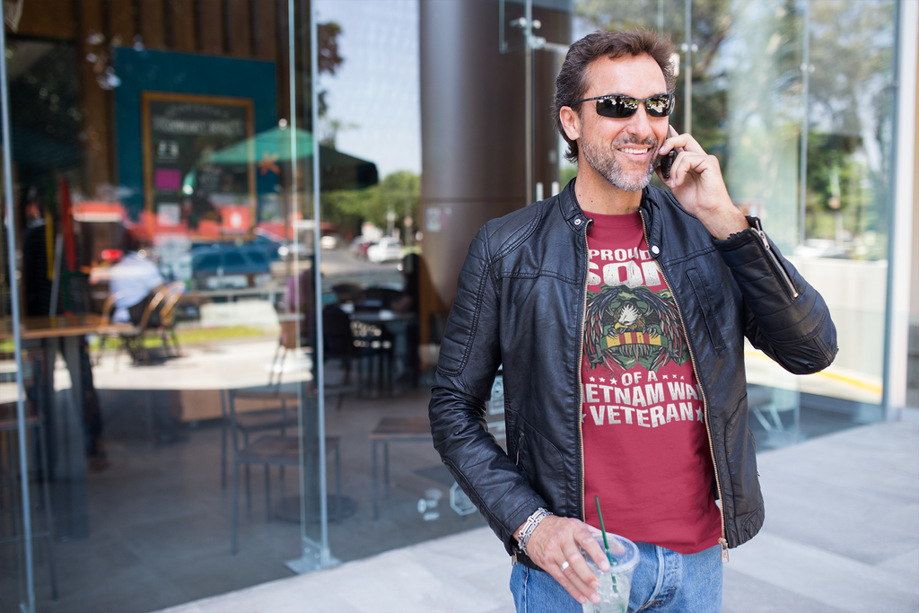 Man wearing Proud Son Of A Vietnam War Veteran shirt, smiling while talking on the phone outside a cafe.