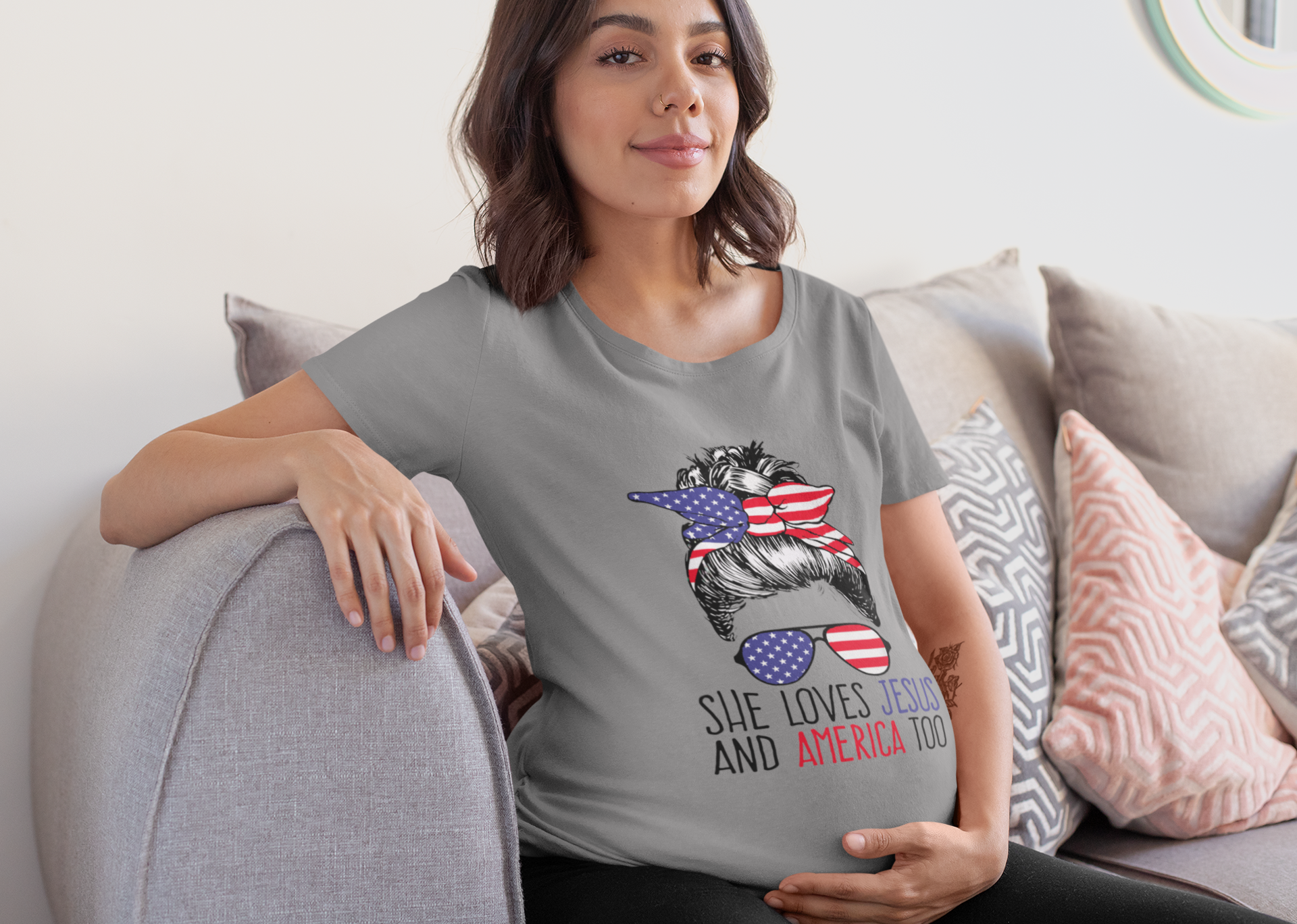 Woman wearing a gray t-shirt with the design "She Loves Jesus And America Too" sitting on a couch.