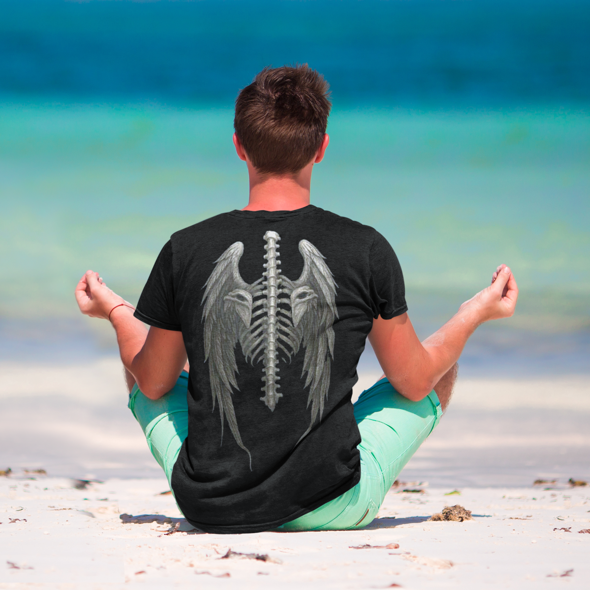 Man meditating on the beach wearing a Spine Eagle Wings Back Print Tee with a unique graphic design.