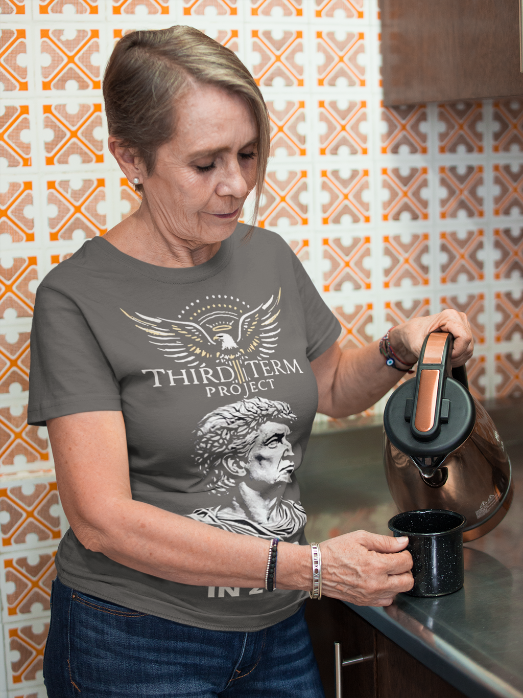Woman pouring coffee while wearing a shirt featuring the Third Term Project Trump 2028 Graphic design in a kitchen.