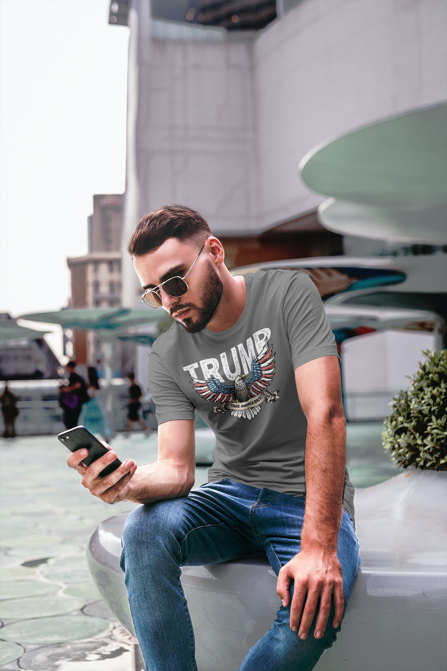 Man wearing a Trump Eagle In Stars And Stripes Take America Back t-shirt while checking his phone in a modern outdoor setting.