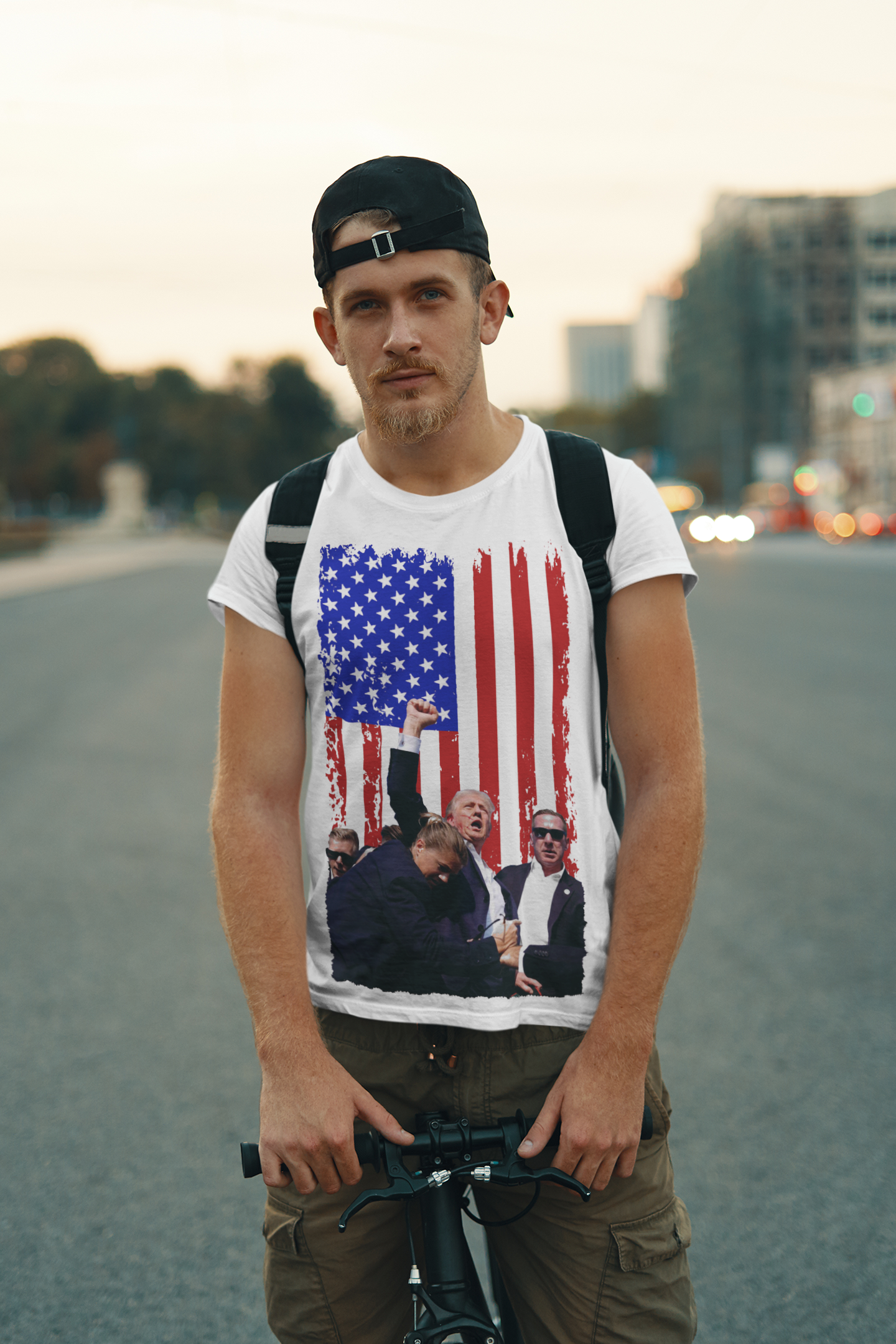 Man wearing a Trump Shot American Flag Design t-shirt, standing with a bicycle on a city street.