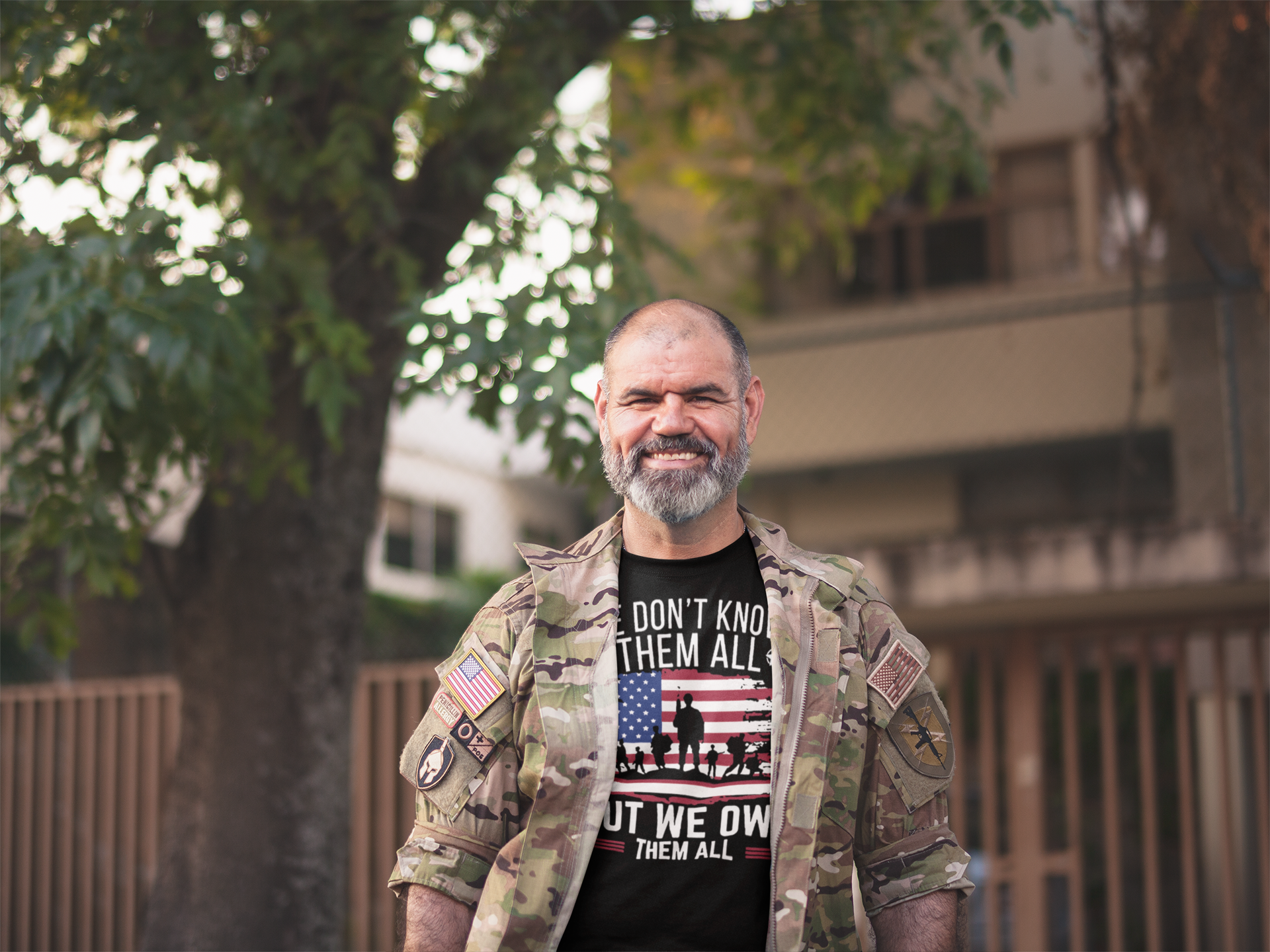 Man in military camouflage wearing 'We Don't Know Them All But We Owe Them All On American Flag' shirt outdoors.
