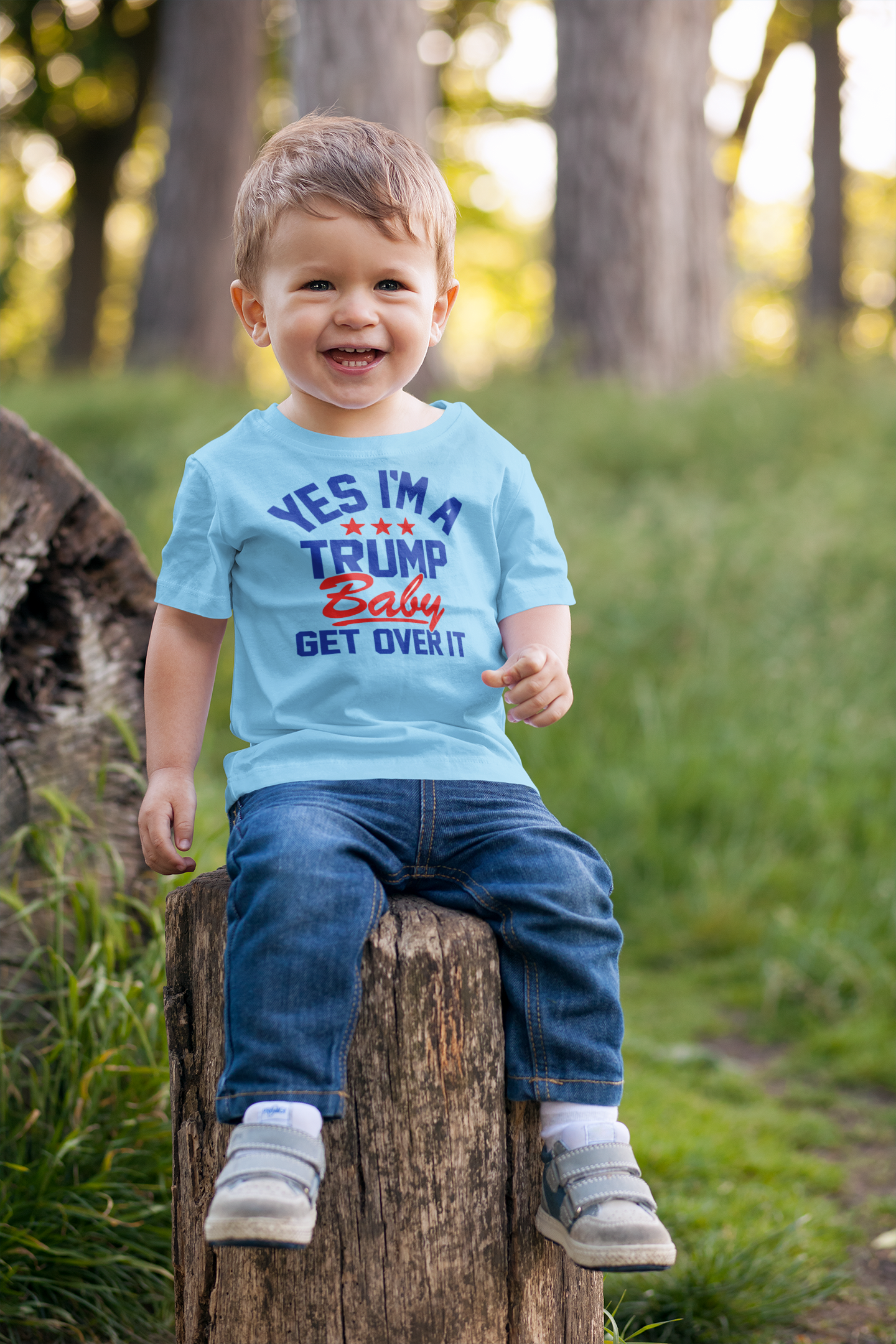 Smiling child wearing a Trump Baby Get Over It Kids Tee while sitting on a log in a sunny outdoor setting.
