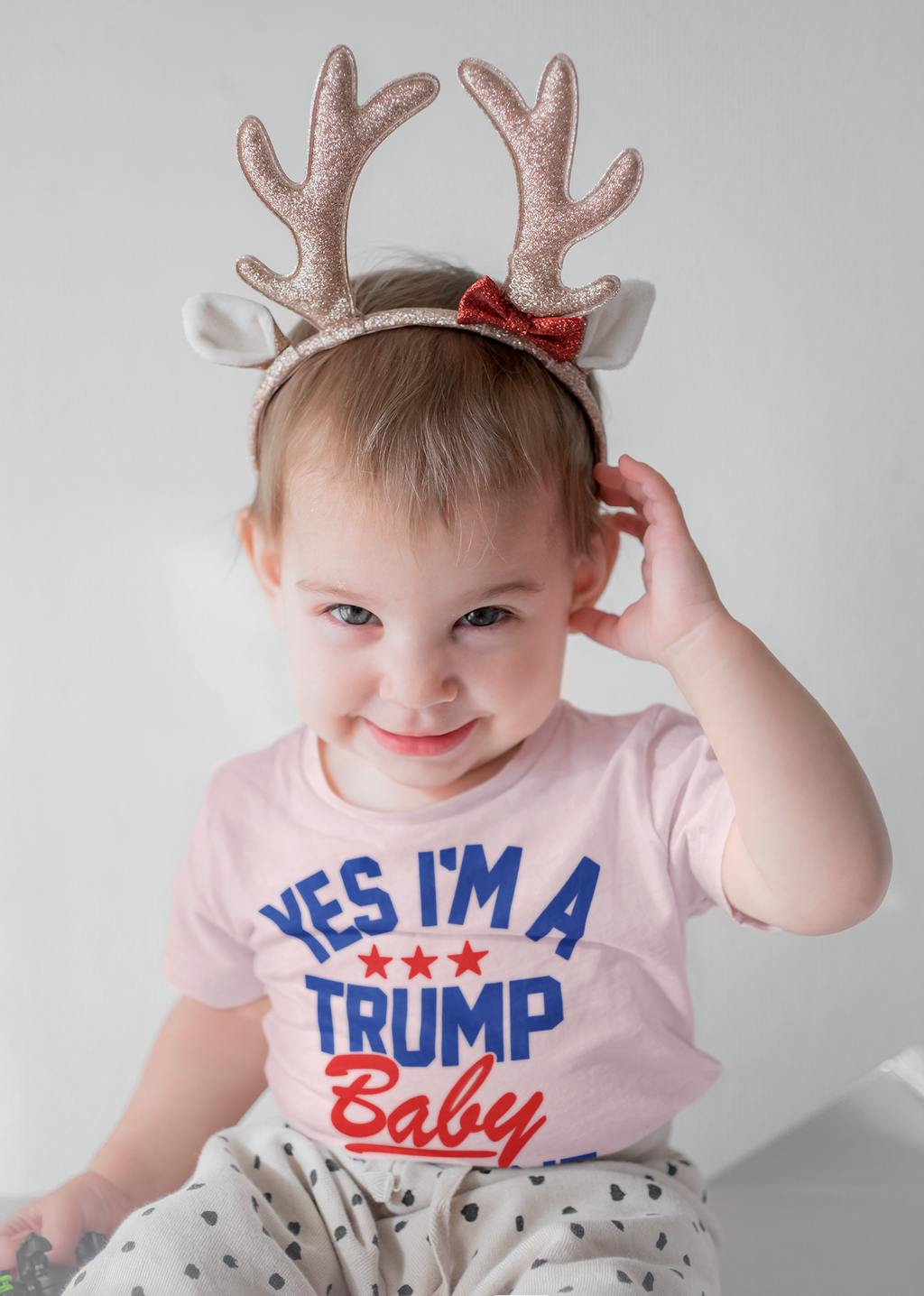 Toddler wearing a Trump Baby Get Over It Kids Tee with festive antlers, smiling joyfully.