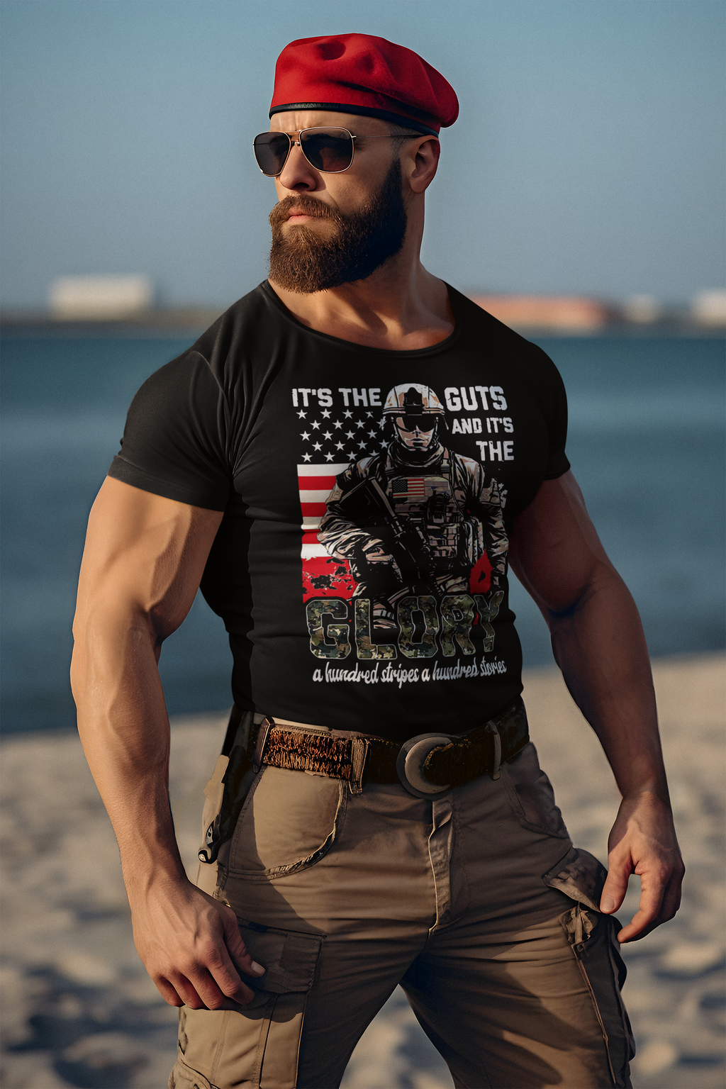 A muscular man wearing a black t-shirt that reads "It's The Guts And It's The Glory" at the beach.