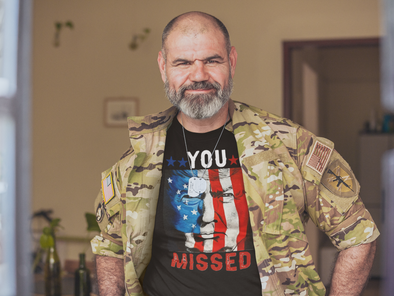 Man wearing a camouflage jacket and t-shirt with a graphic design, standing indoors.