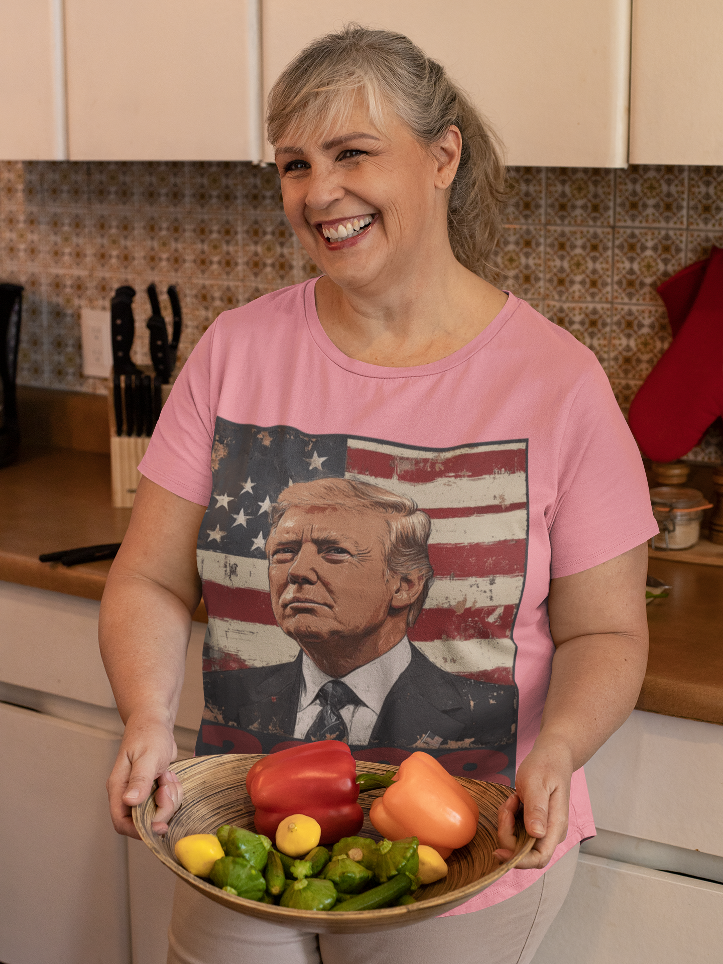 Woman smiling in a kitchen wearing Trump 2028 USA Flag Motif Gear t-shirt and holding a bowl of fresh vegetables.