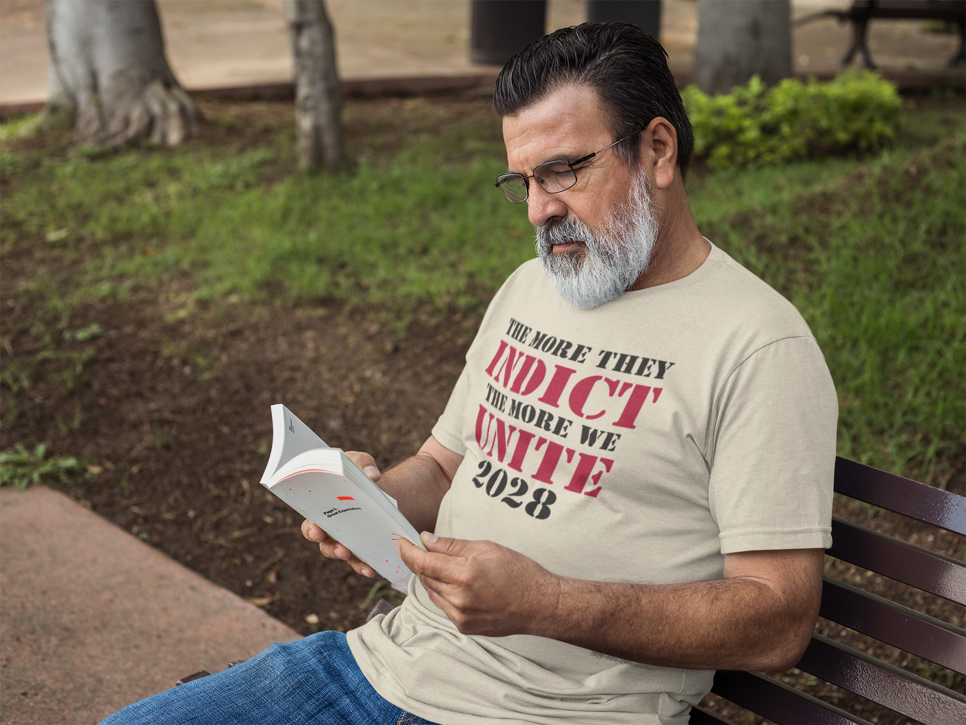 A man reading a pamphlet while wearing a shirt that says The More They Indict The More We Unite 2028 Blk Ltrs.