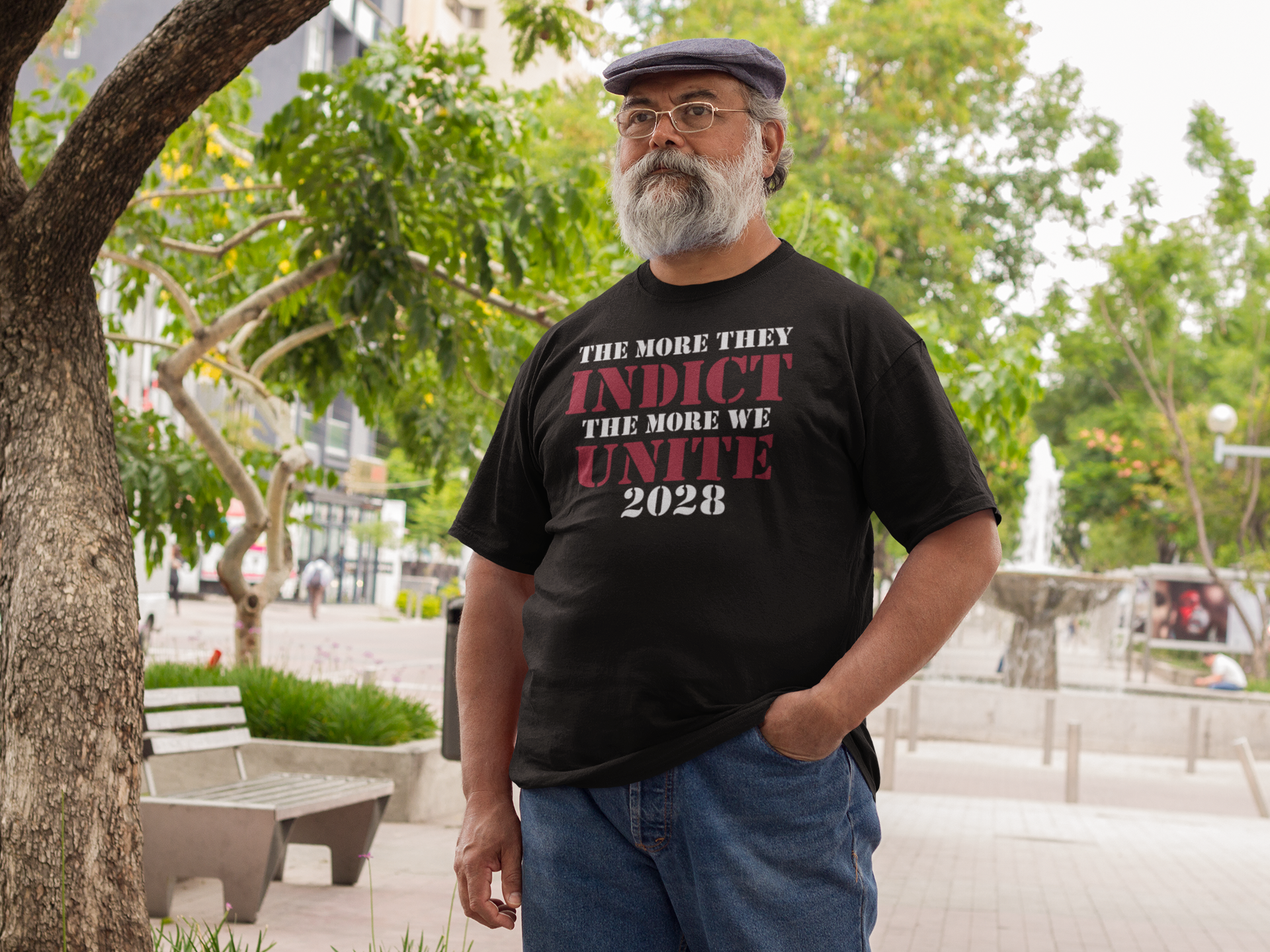 Man wearing a black t-shirt with the phrase The More They Indict The More We Unite 2028 Wht Ltrs in a park.