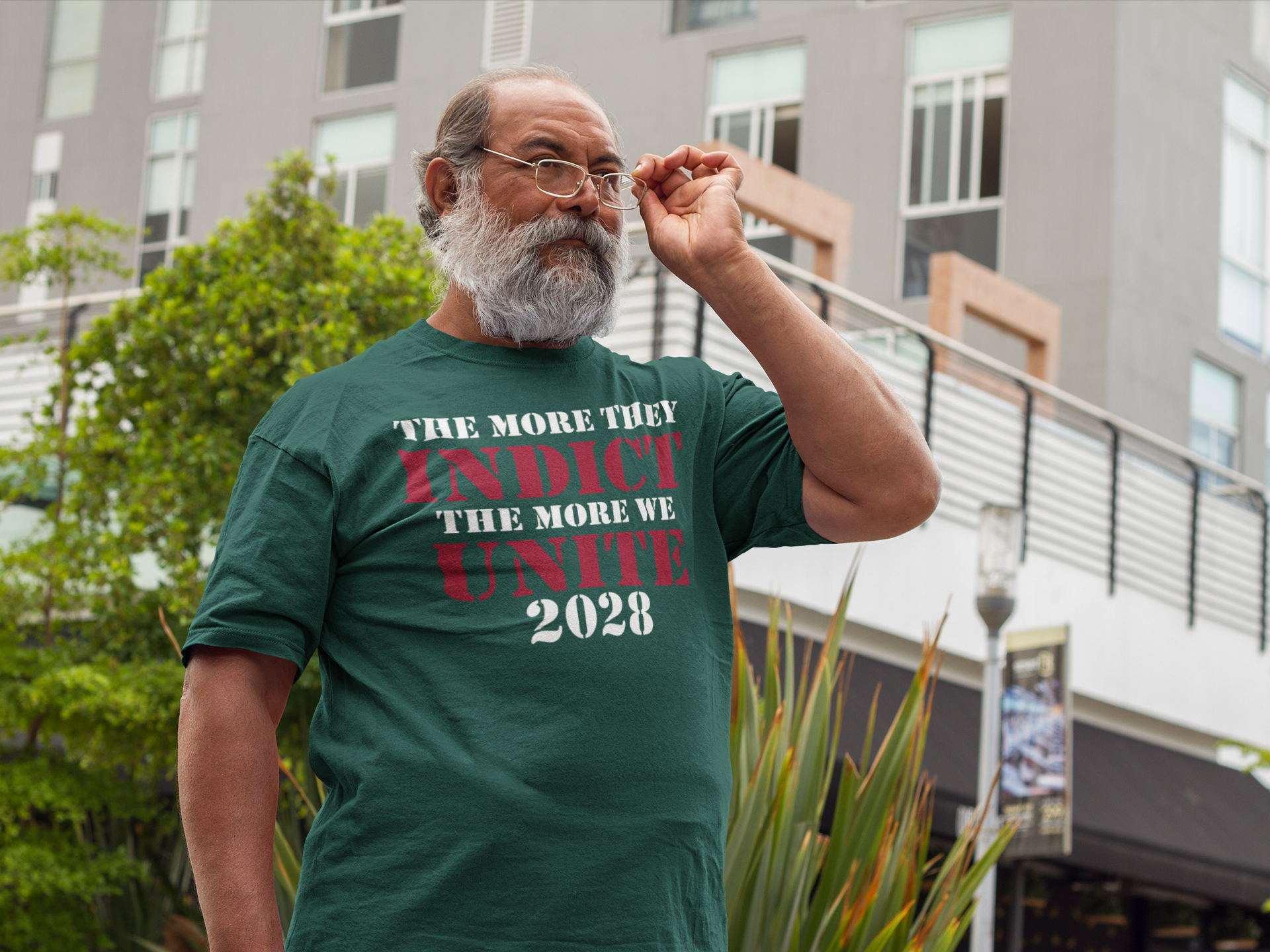 The More They Indict The More We Unite 2028 Wht Ltrs on a green t-shirt worn by a man outdoors.
