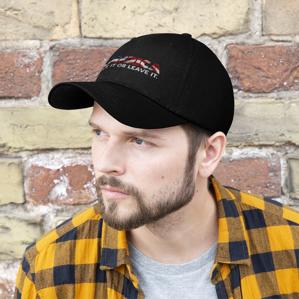Man wearing America Love It Or Leave It Twill Baseball Cap against a brick wall, showcasing a patriotic statement.