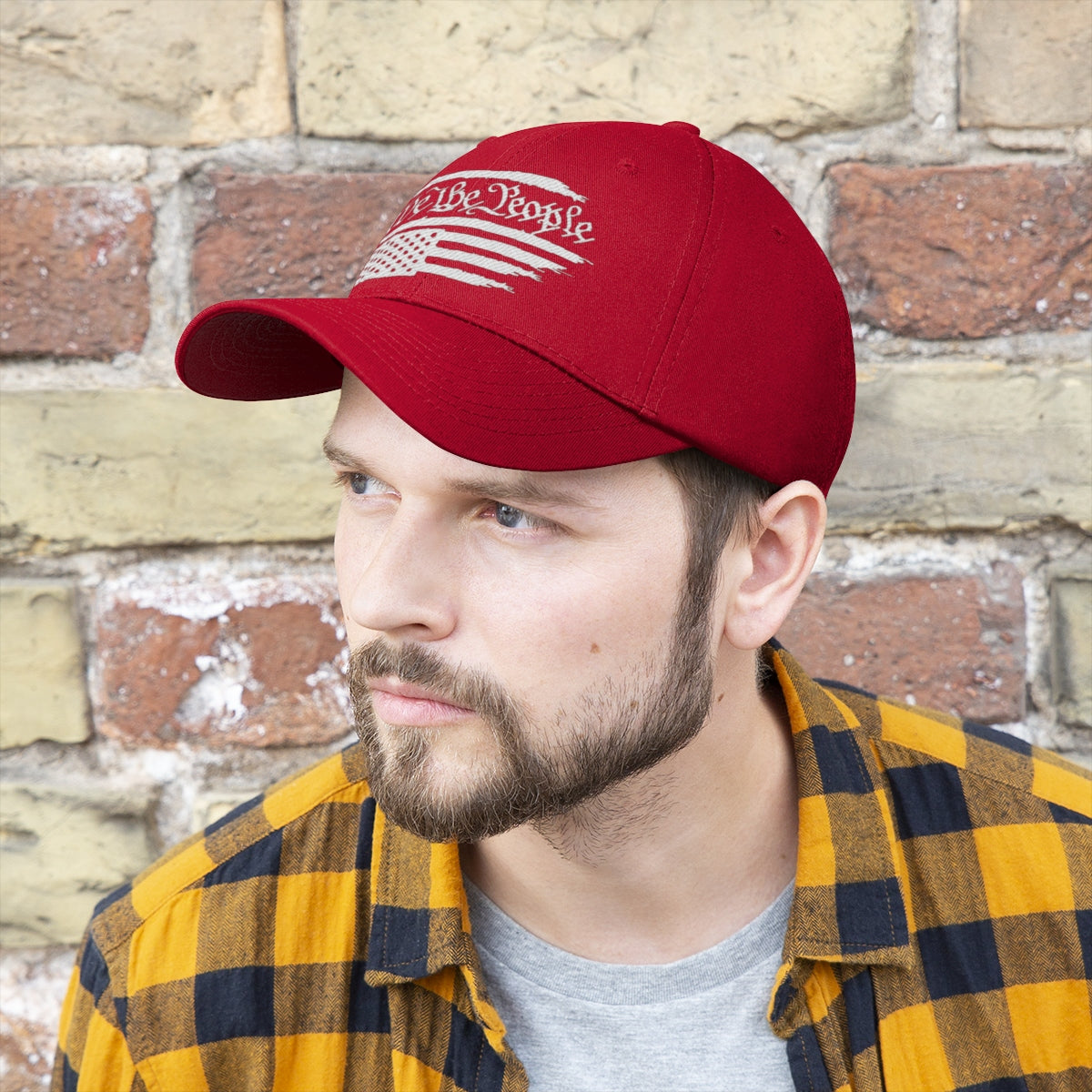 We The People On Distressed American Flag Twill Cap worn by a man with a beard, standing against a brick wall.