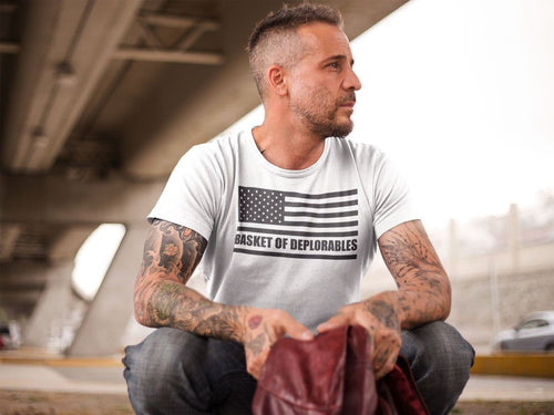 Man wearing Basket Of Deplorables On American Flag shirt with tattoos on arms, sitting under a bridge.