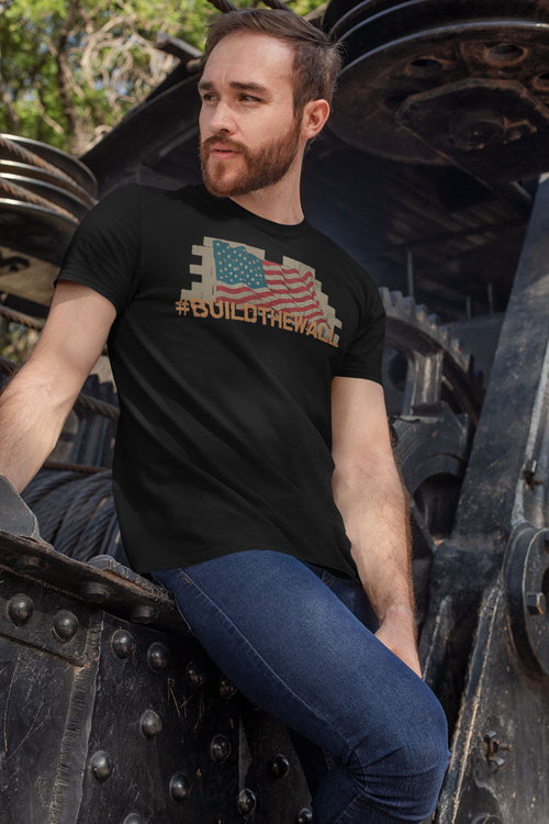 Man wearing a black t-shirt with 'Build The Wall Bold Statement Design' in front of industrial background