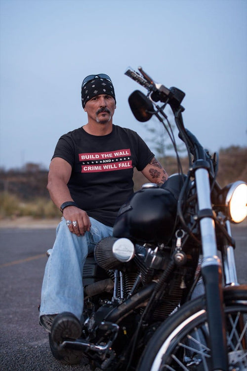 Man in 'Build The Wall And Crime Will Fall' t-shirt sitting on a motorcycle, showcasing bold support for national border security.