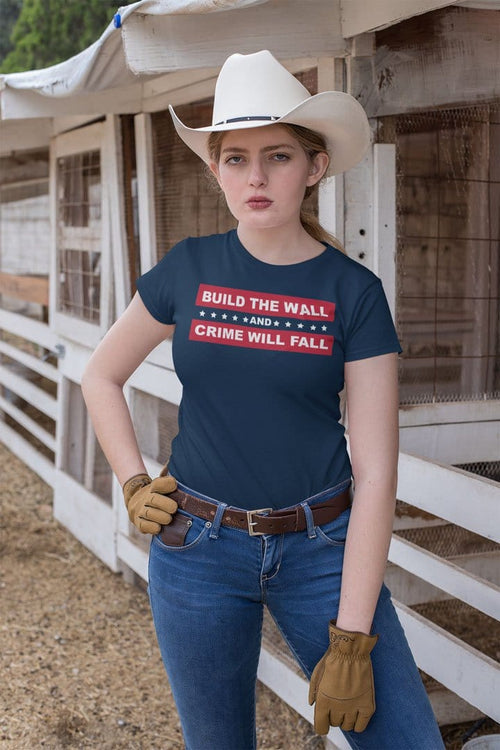 Woman wearing 'Build The Wall And Crime Will Fall' t-shirt, cowboy hat, and jeans, standing by a rustic fence.