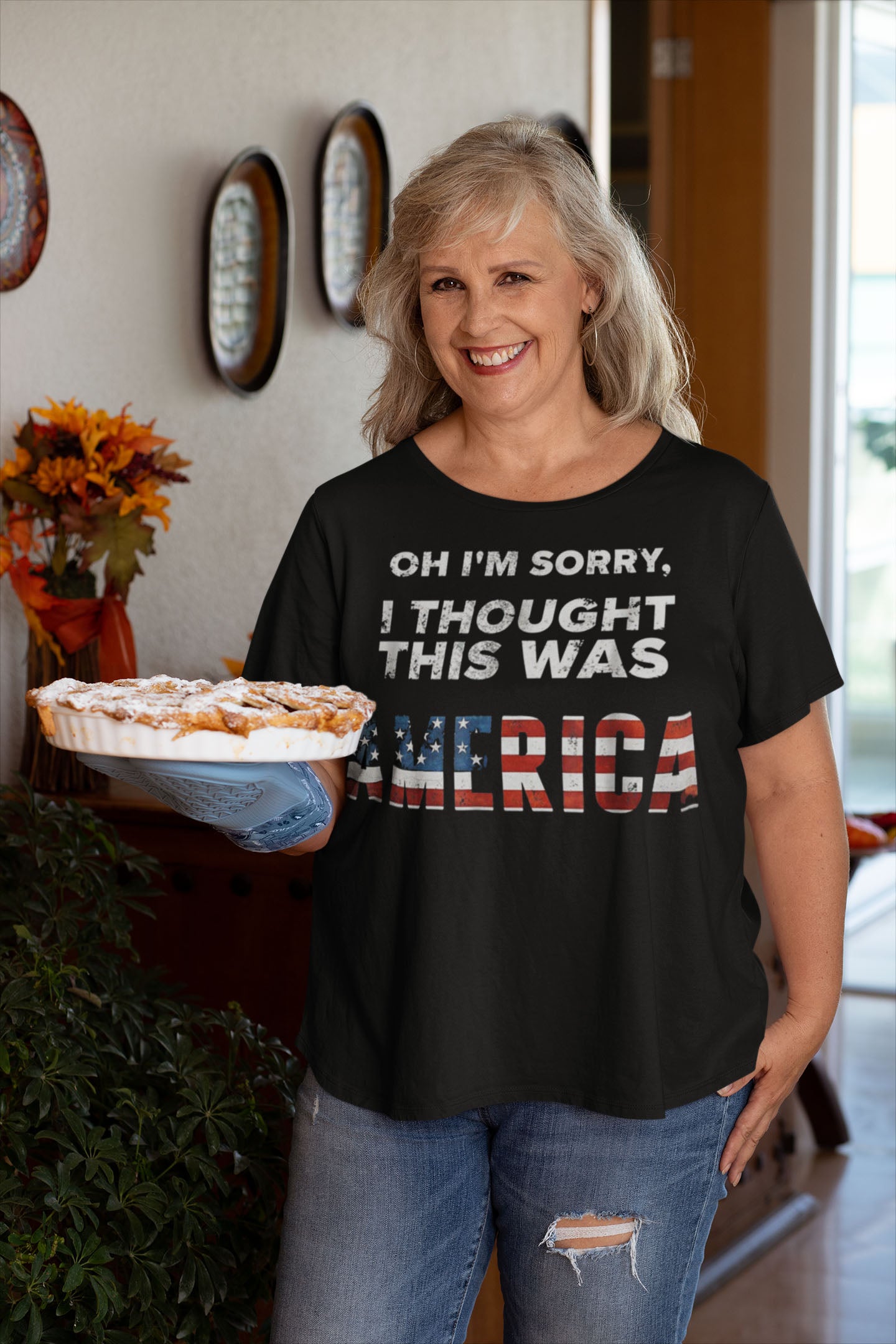 Woman wearing 'Oh I'm Sorry I Thought This Was America' shirt holding pie indoors.