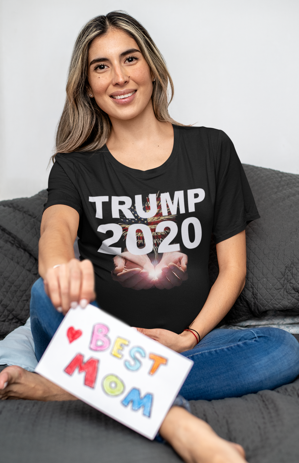 Woman wearing Trump 2020 Heavenly Hands Patriotic Tee holding a 'Best Mom' sign while sitting on a couch.