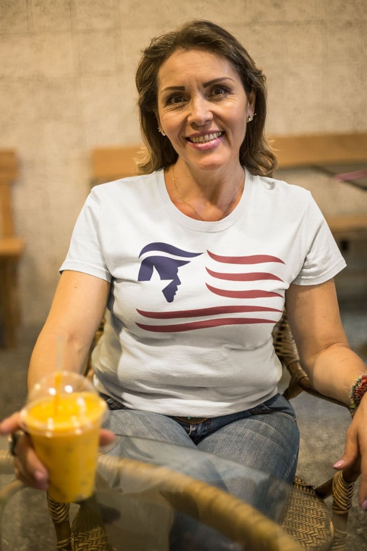 Woman wearing a shirt with Trump Silhouette As American Flag design while sitting and holding a drink.