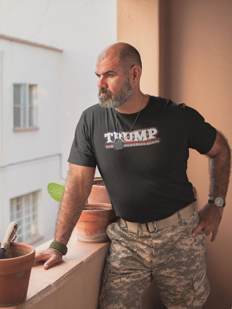 Man wearing Trump On American Flag Make America Great Again shirt on a porch, military-style pants, looking into distance.