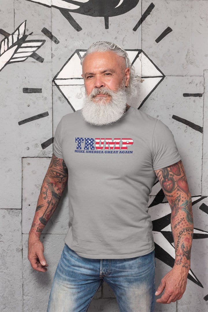 Man wearing Trump As American Flag Make America Great Again t-shirt with tattoos and a white beard, standing against a wall.
