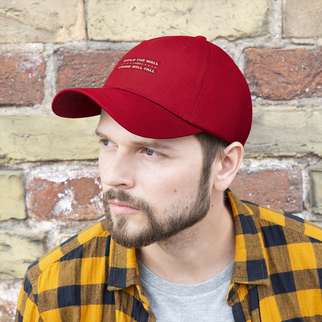 Build The Wall And Crime Will Fall Twill Cap worn by a man in a plaid shirt against a stone wall.