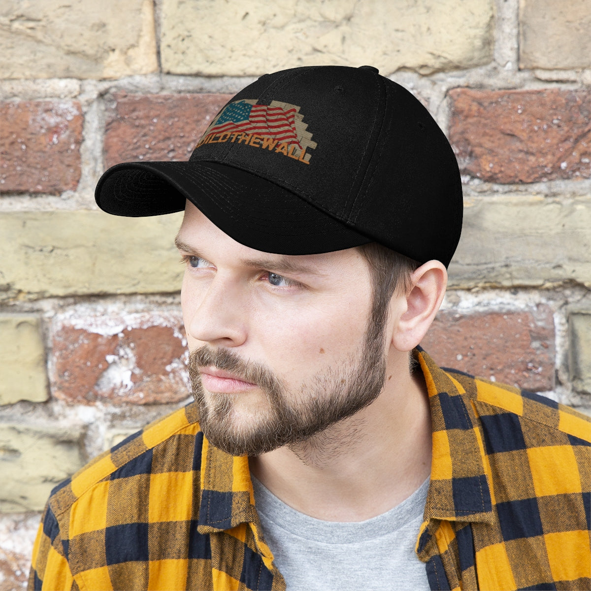 Build The Wall On American Flag Twill Cap worn by a man in front of a brick wall, showcasing its patriotic design.