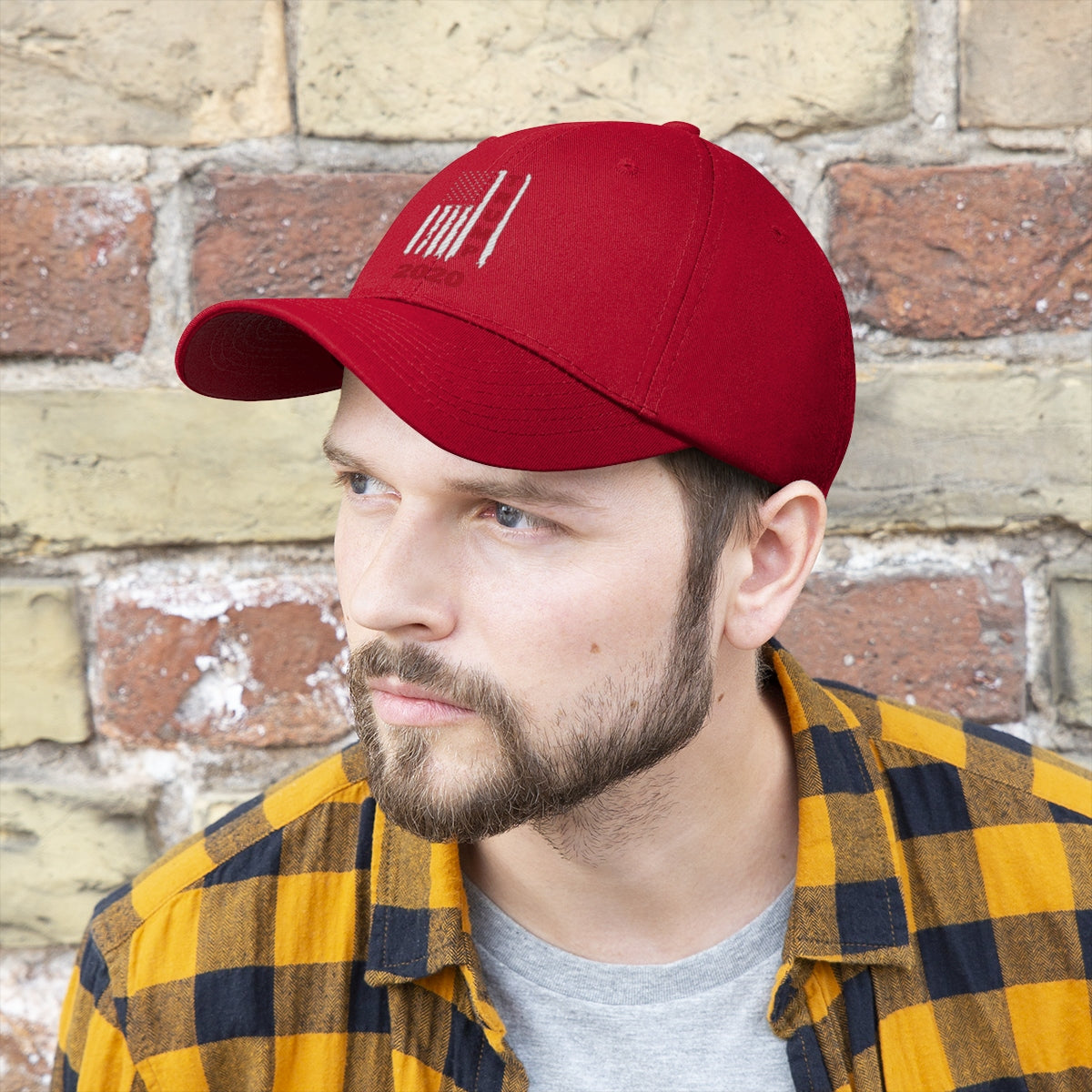 Trump 2020 Red On Distressed American Flag Twill Cap worn by a man in front of a brick wall.