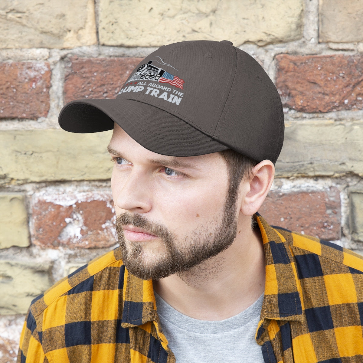 Man wearing All Aboard The Trump Train 2020 Twill Cap against a brick wall, showcasing political support.
